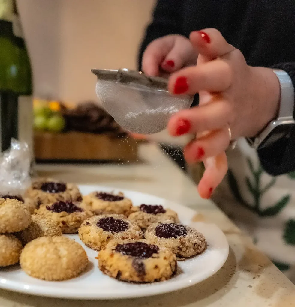 Visual of the baker dusting powdered sugar over jam thumbprint cookies, made from Vanilla Sugar Cookie dough.