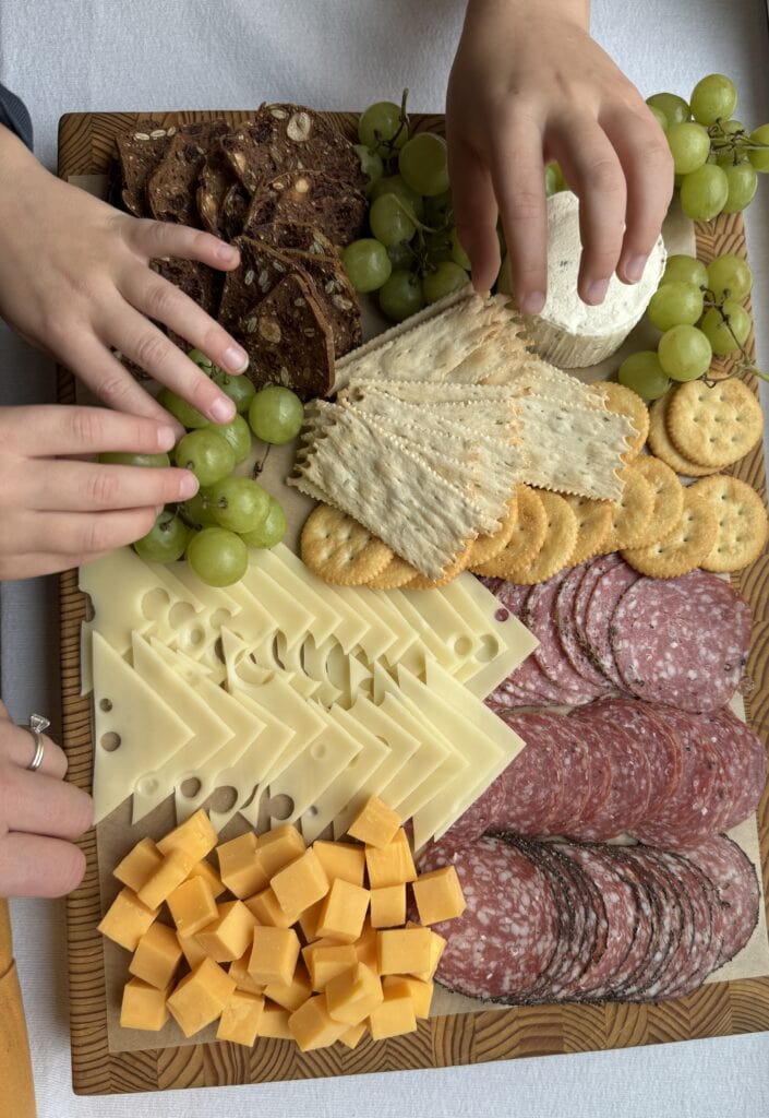 Cheese and salami board with crackers and grapes; Guests' hands reaching in to grab something to eat.