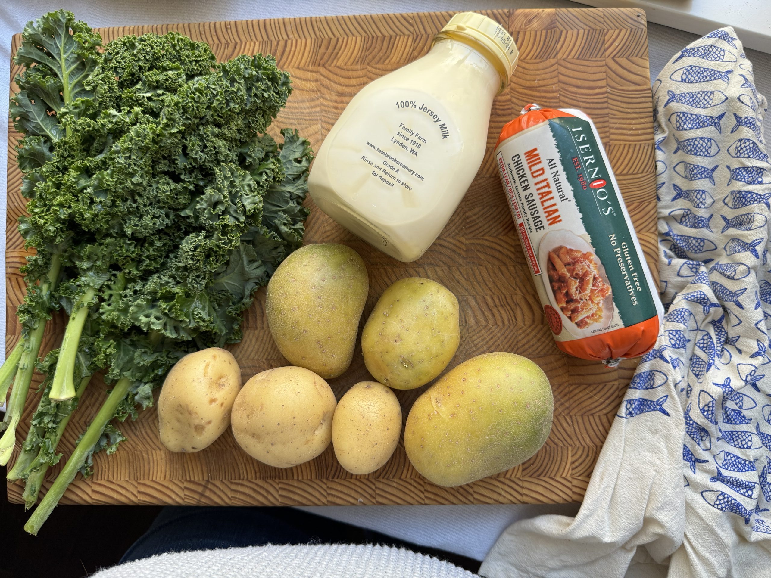 Overhead image of ingredients, prior to preparing the dish. A bunch of kale, ping of cream, Yukon Gold potatoes, and a roll of Italian chicken sausage.