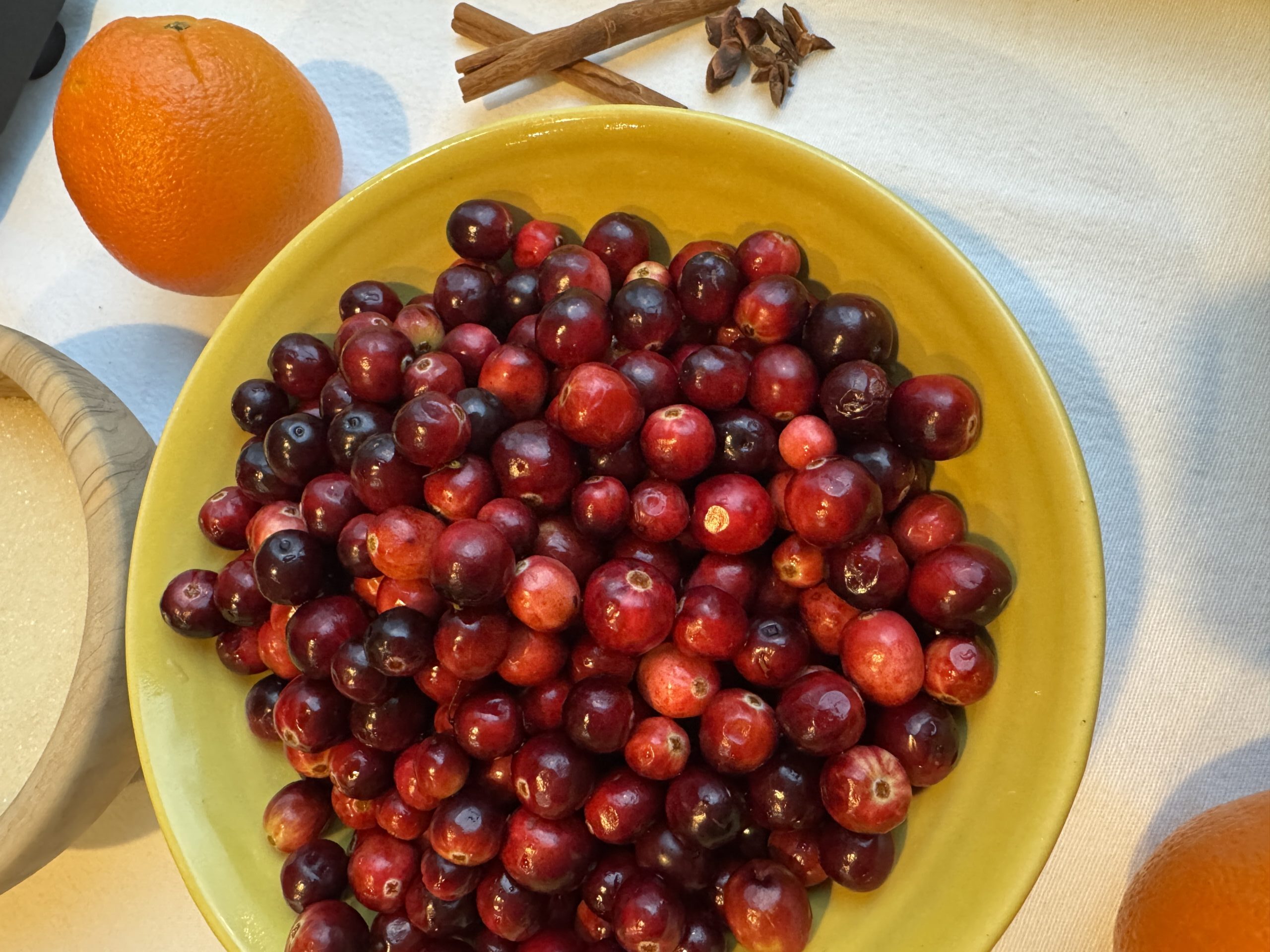Fresh cranberries in a yellow ceramic bowl, an orange, cinnamon sticks, star anise, and sugar.
