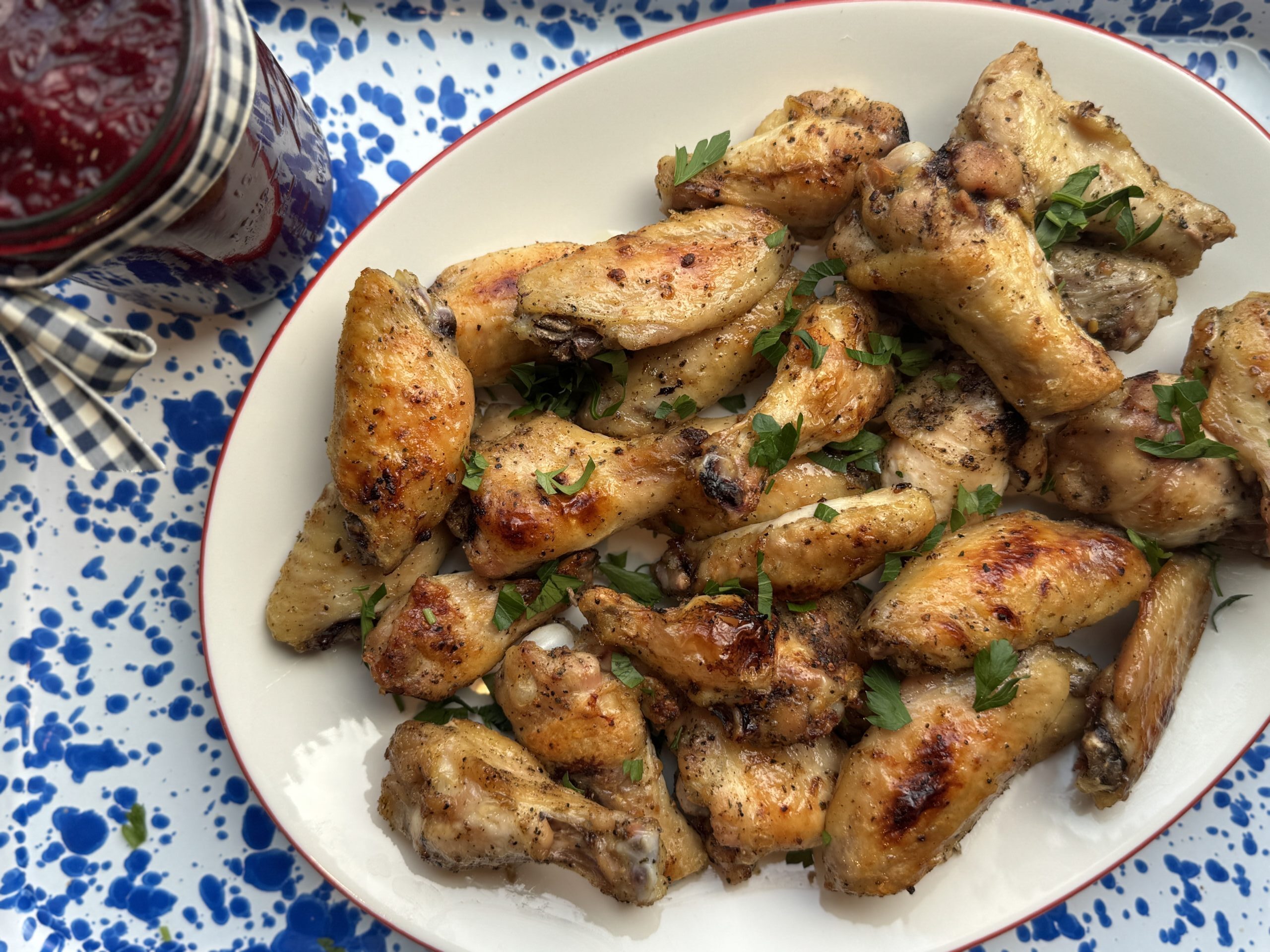 Overhead image of finished product: golden brown Dry Rub Chicken Wings topped with chopped Italian parsley. The Wings are on a white oval platter with red trim; a small jar of cranberry sauce is placed just to the right of the platter.