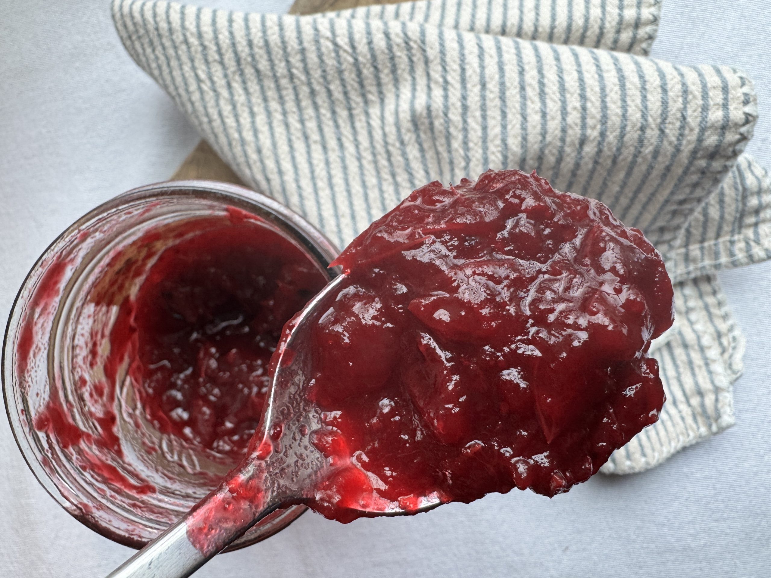 Finished cranberry sauce in a jar being spooned out. Striped napkin to the side.