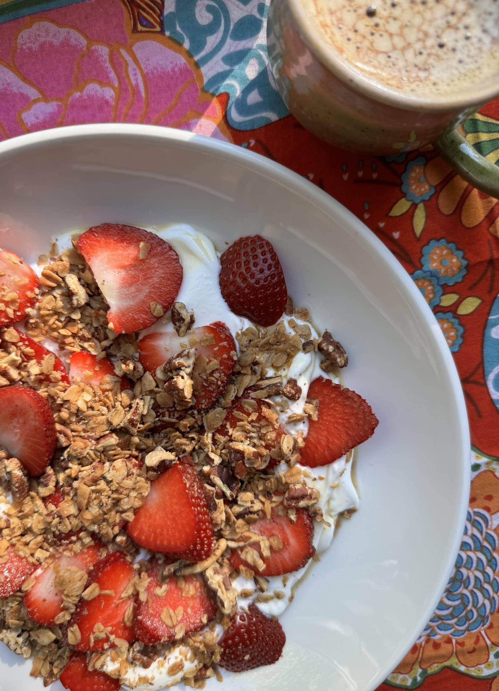 Finished product, Gluten-Free Granola, with strawberries in a bowl. A mug of hot coffee next to the bowl.