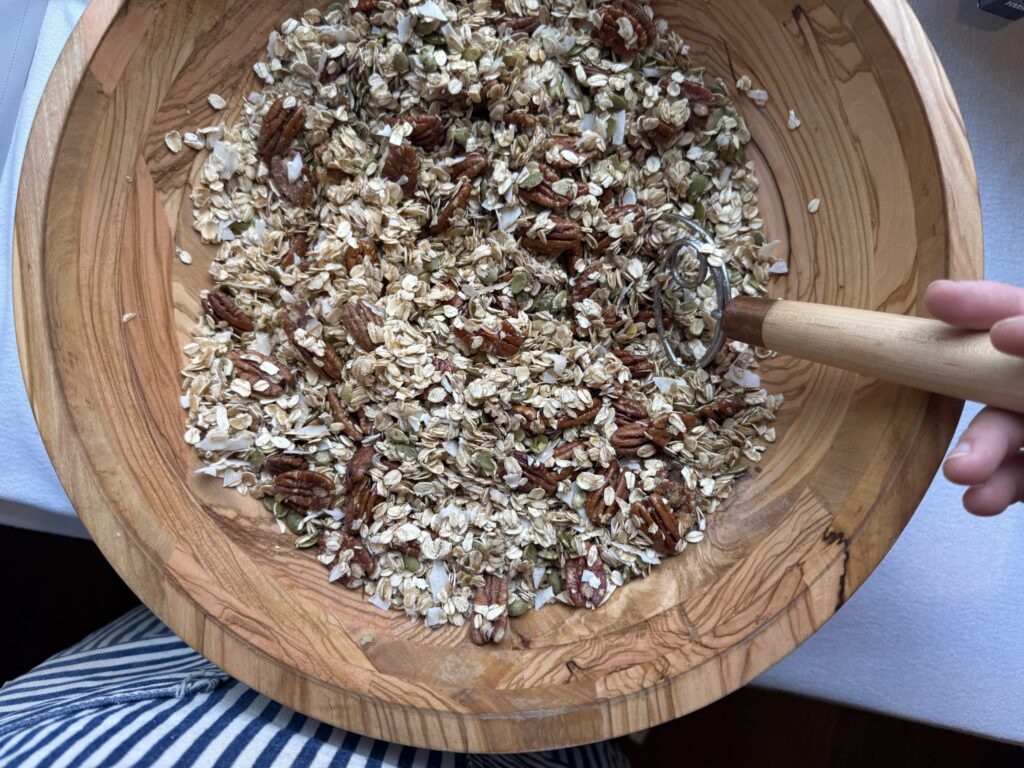 Raw ingredients for Gluten-Free Granola in a wooden bowl, a hand stirring with a Danish whisk.