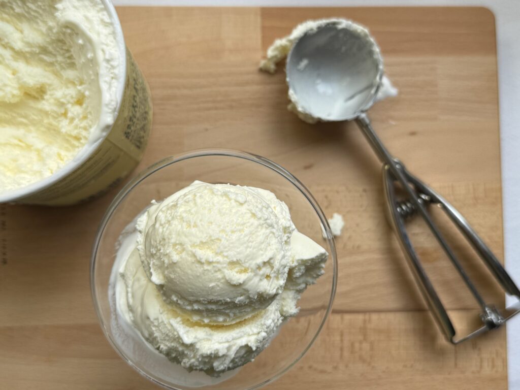 Vanilla ice cream in the carton, alongside a scoop in a clear glass, alongside a scoop.