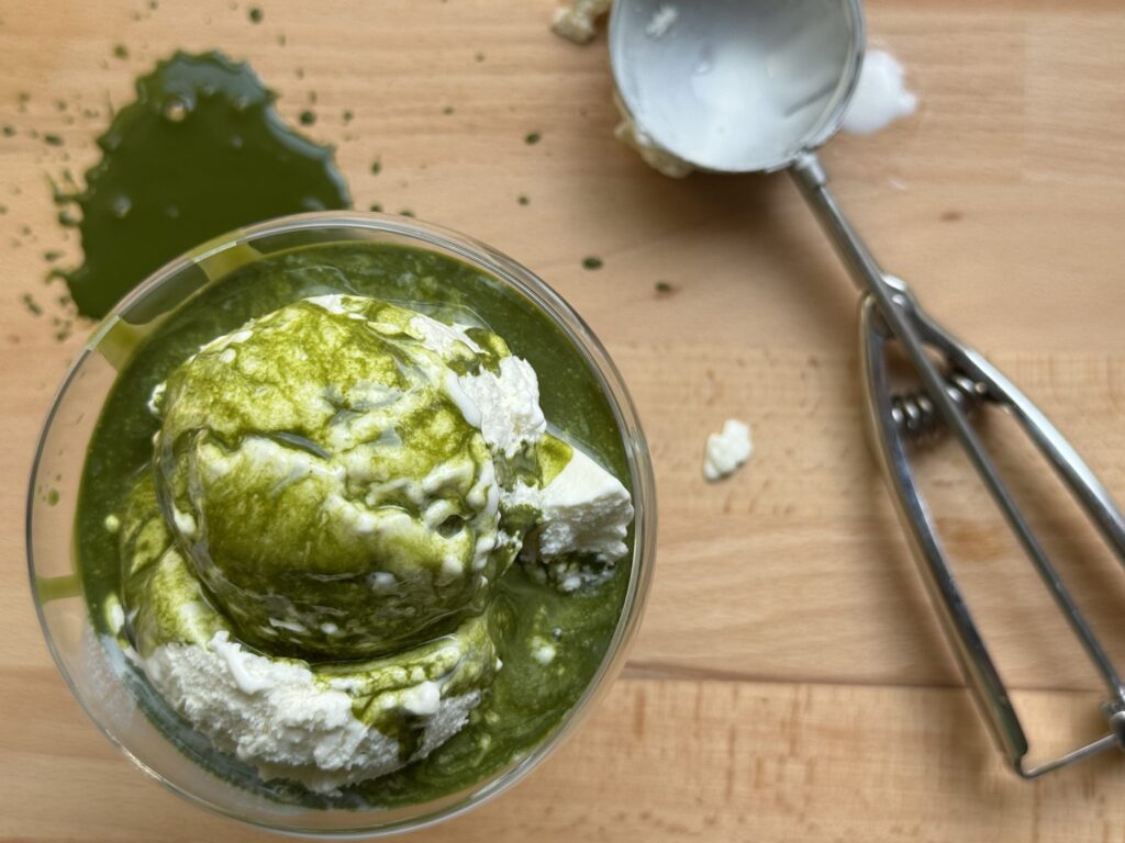 Matcha green tea poured over a scoop of vanilla ice cream. There's an ice cream scoop to the right of the dessert.