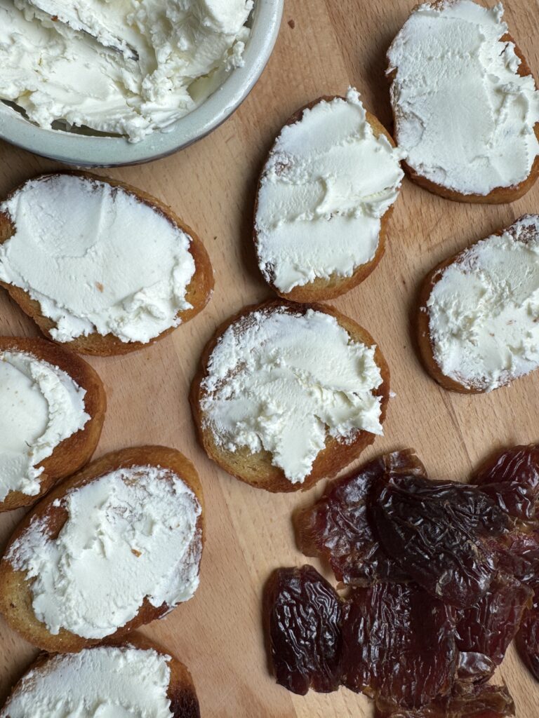 Crostini with roughly spread goat cheese on top; dates on the cutting board next to the crostini.