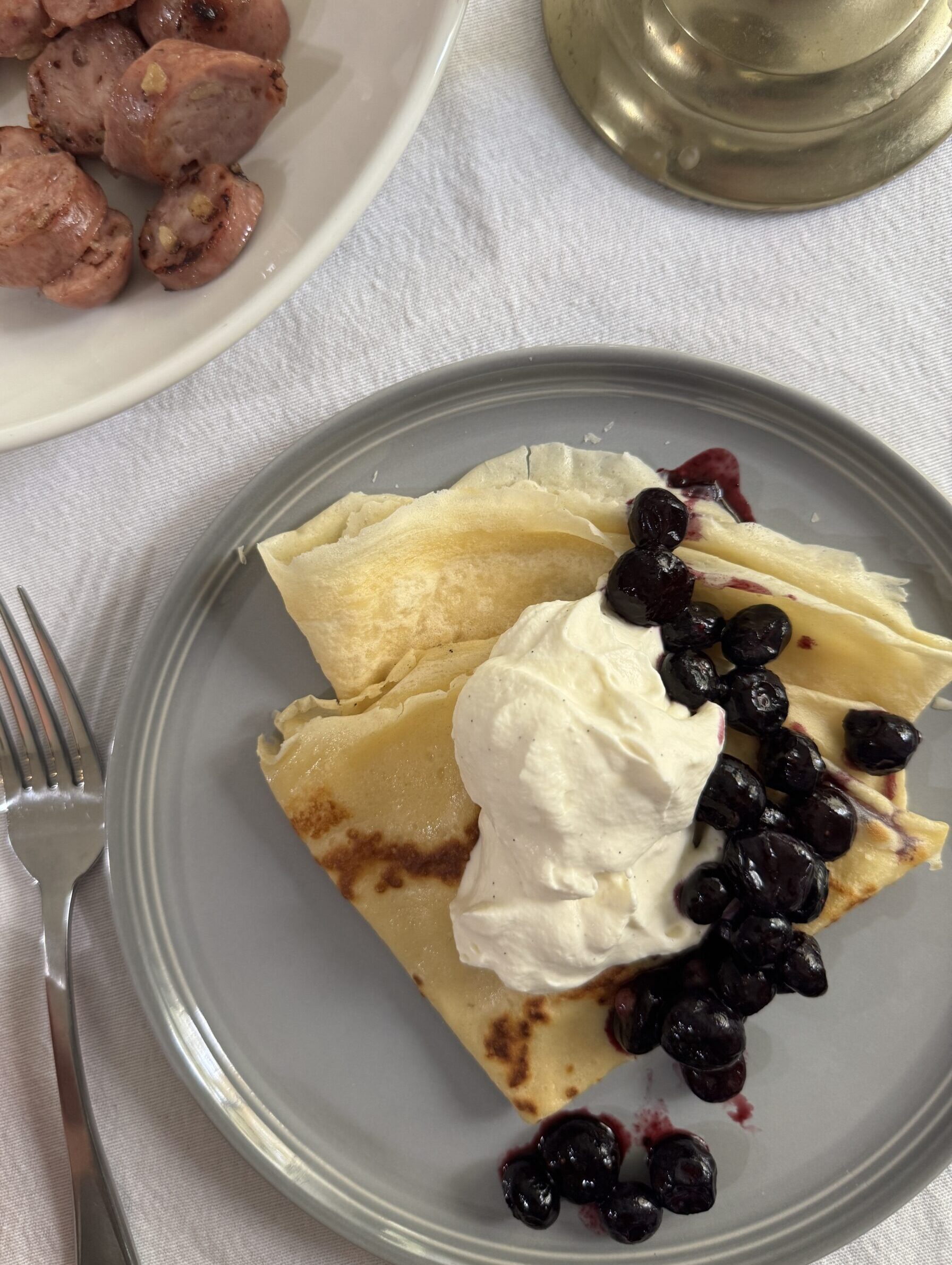 Two crepes, perfect for one person, served with vanilla bean whipped cream, blueberry compote, and a side of sausage.