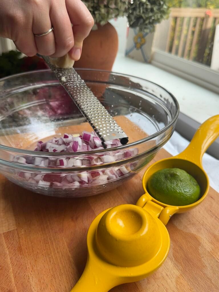 Person grating red onion into a glass mixing bowl for fresh salsa, with a yellow lemon juicer nearby.