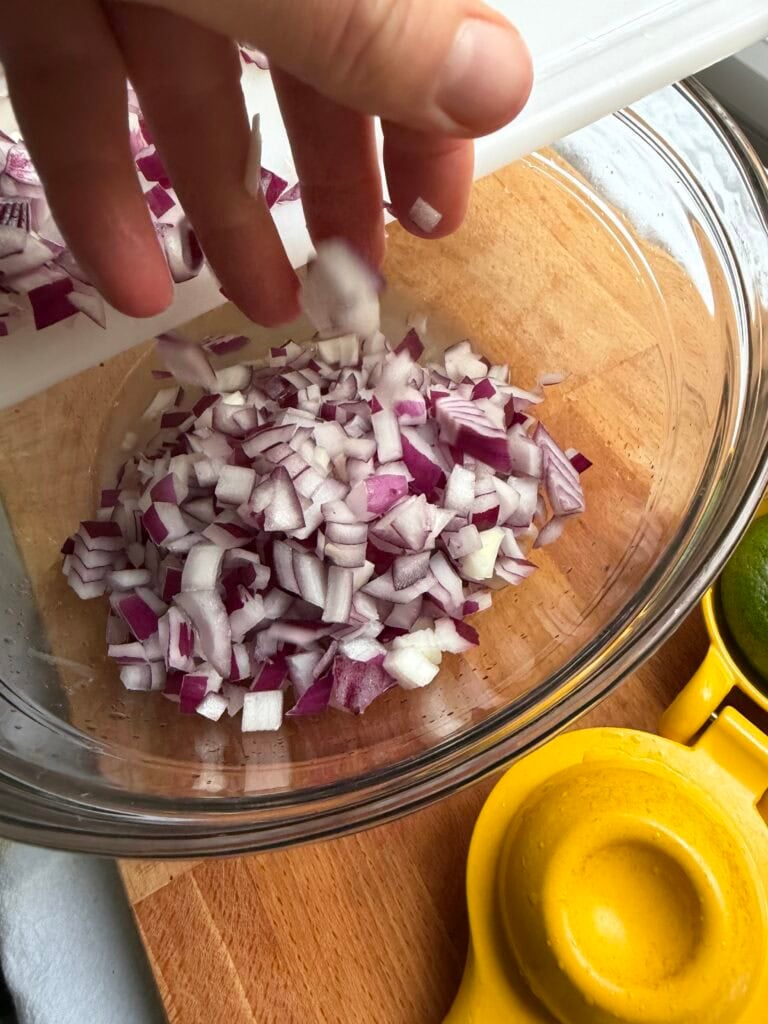 Adding diced red onions to a clear bowl with lime juice for black bean and corn salsa.